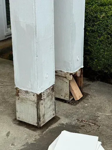 White porch posts with damaged wood bases and a loose board on concrete near a hedge.