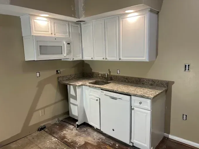 Small kitchen with white cabinets, granite countertop, and a microwave, showing unfinished flooring and bare walls