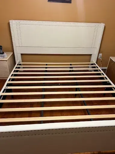White upholstered bed frame with wooden slats in a bedroom, shown without a mattress.