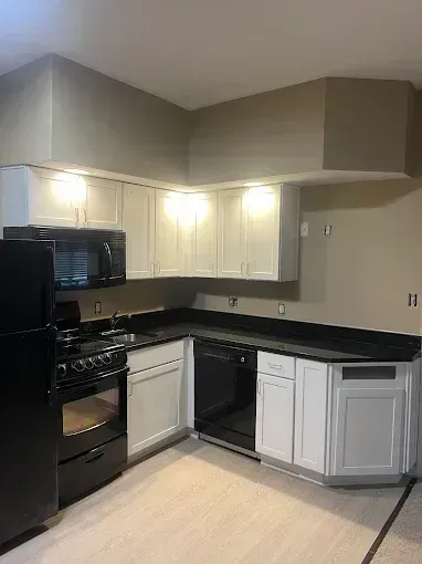 Compact kitchen with black appliances, white cabinets, and black countertops under a sloped ceiling