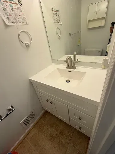 Small white bathroom vanity with sink and mirror, chrome faucet, towel ring, and tan tile floor.