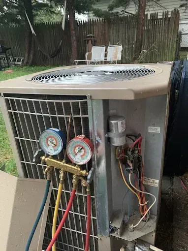 Outdoor HVAC unit with exposed gauges and wiring, sitting beside a fence in a yard