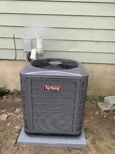Gray AirTemp outdoor air conditioner unit on a concrete pad against green siding.