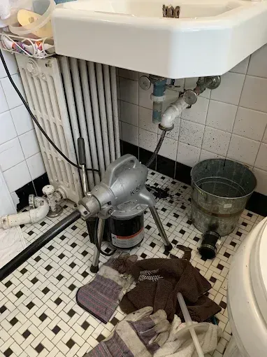 Cluttered bathroom with broken sink plumbing, bucket, and towels on a tiled floor.