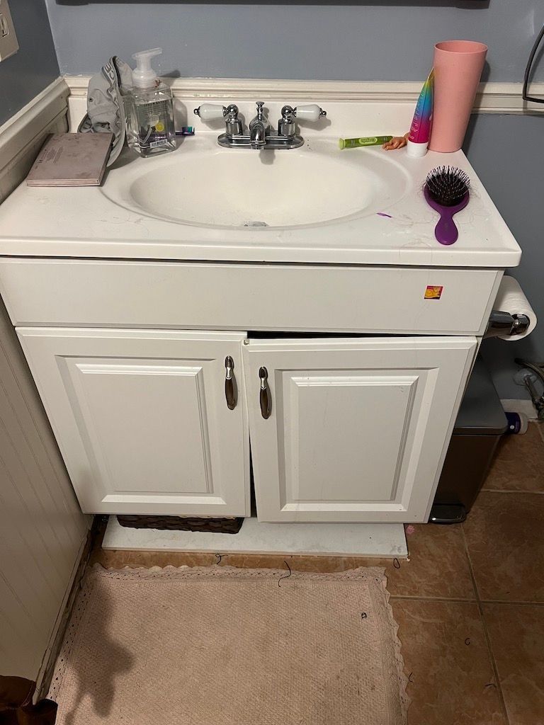 White bathroom vanity with sink, faucet, toiletries, and beige rug on tiled floor