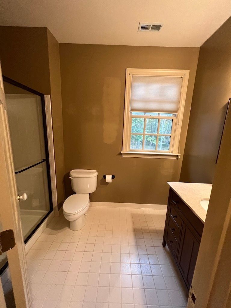 Bathroom with beige walls, white tile floor, toilet, shower, window, and dark wood vanity.