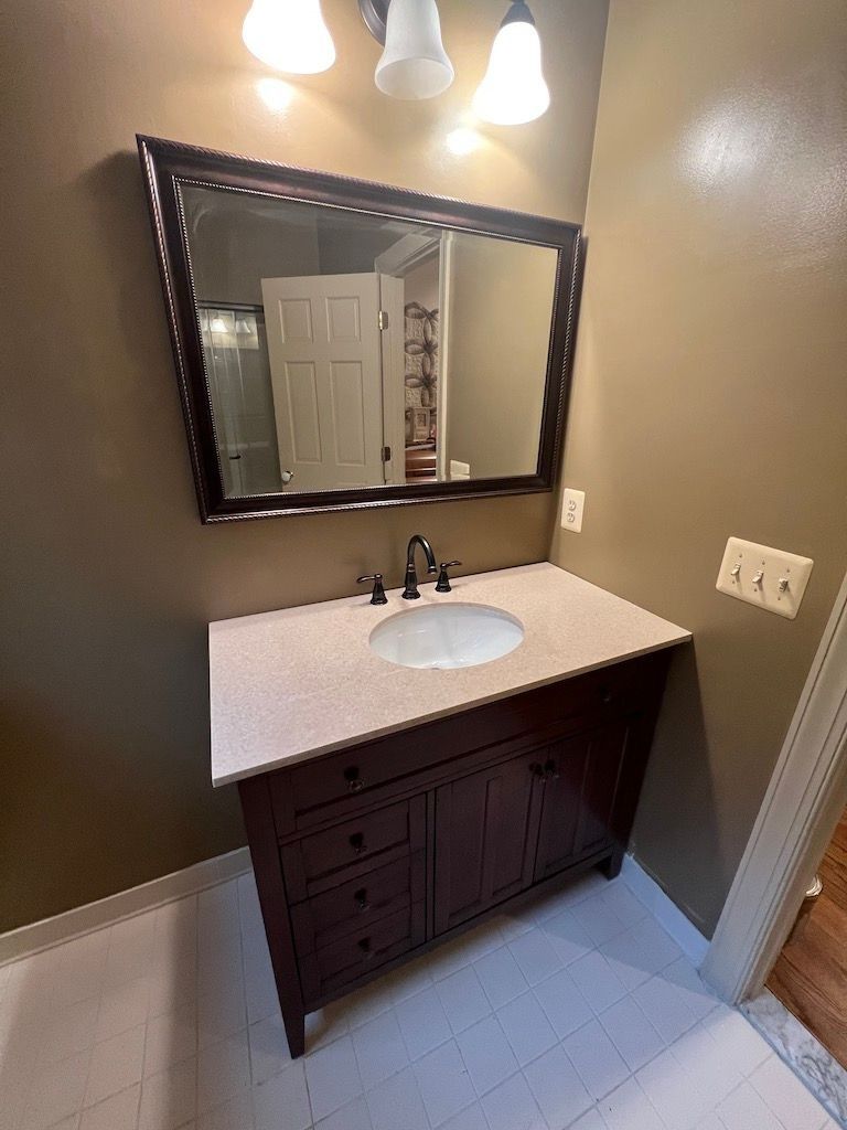 Bathroom vanity with dark wood cabinet, white countertop, round sink, mirror, and three lights on beige walls