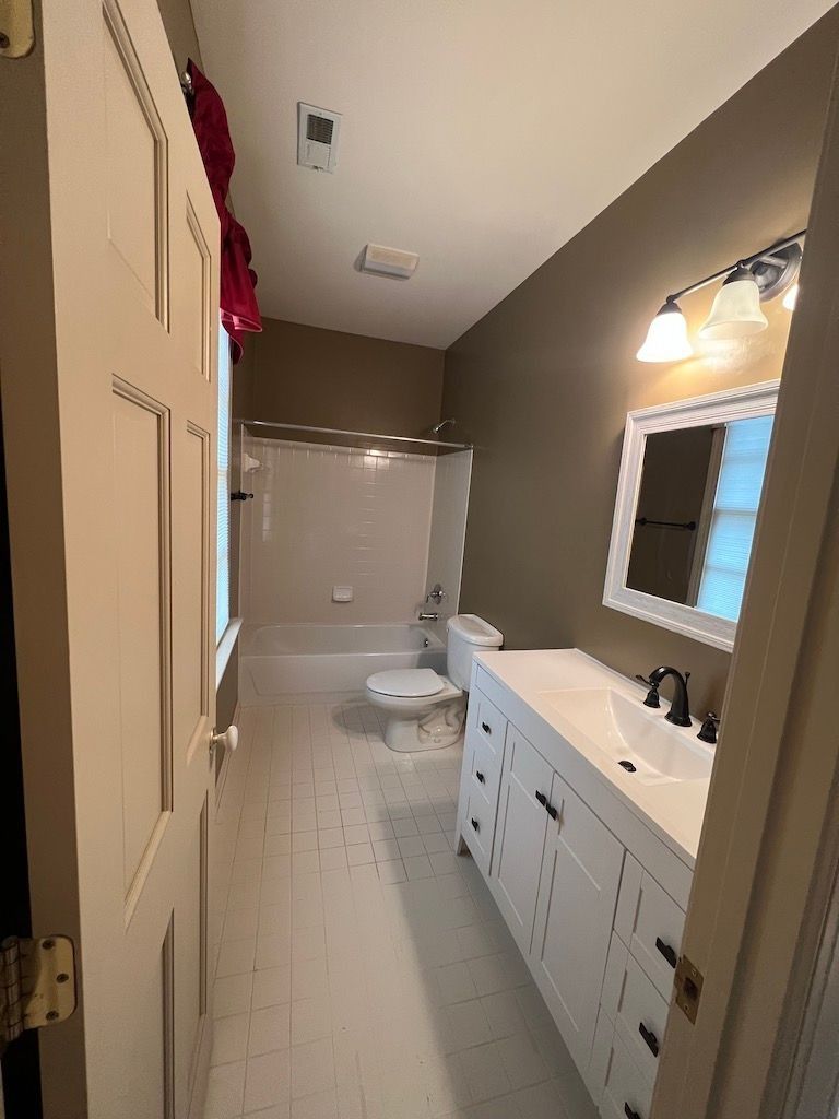 Narrow bathroom with beige walls, white vanity, toilet, shower curtain, and sink under a lit mirror.