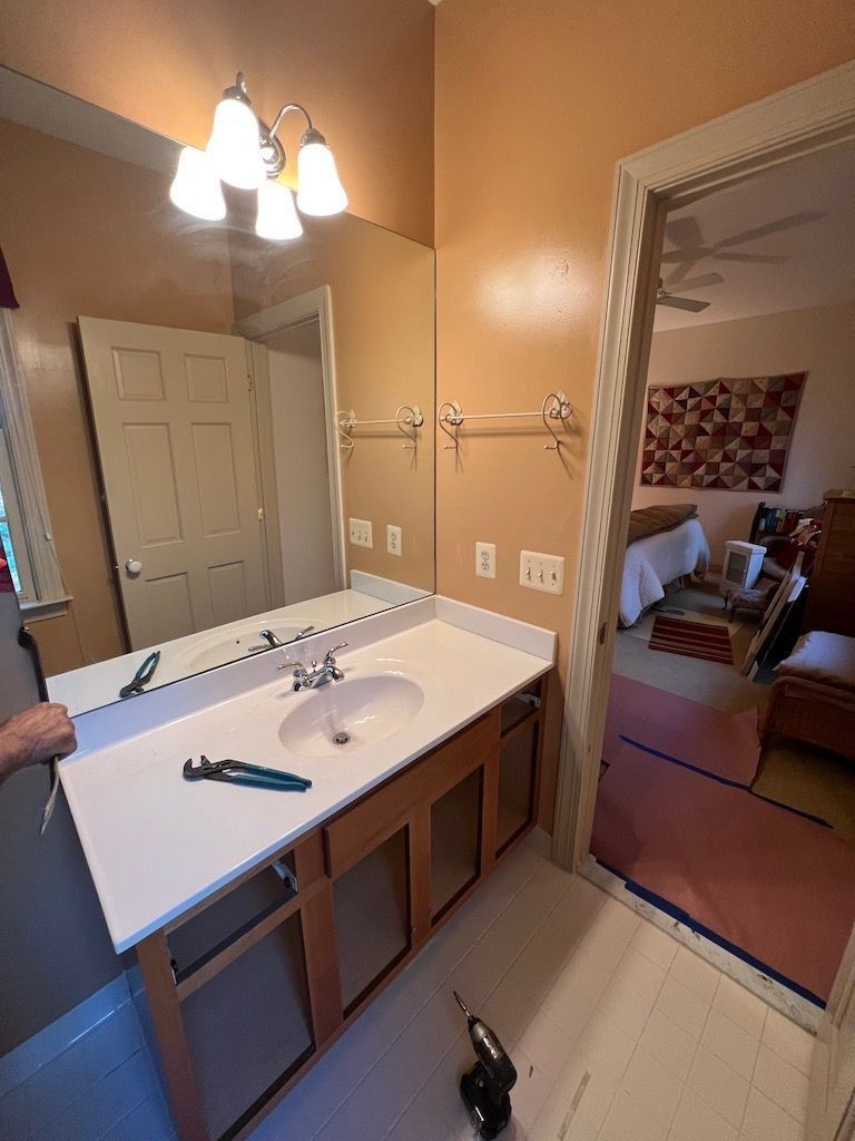 Bathroom vanity with double sinks and mirror, viewed through an open door into a bedroom.