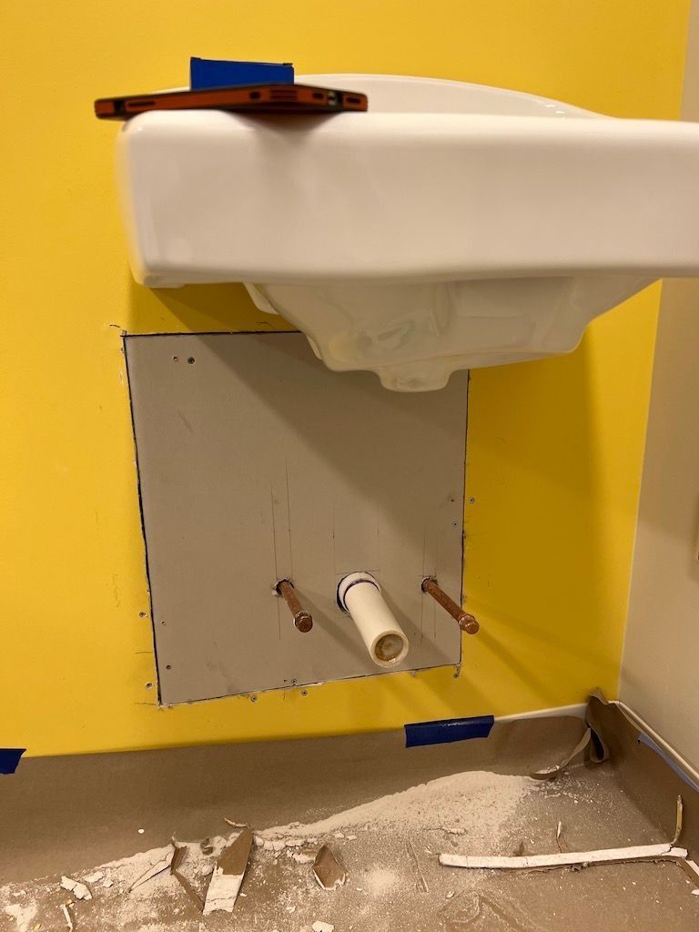 Sink mounted on a yellow wall above a rough wall opening with exposed plumbing and a white pipe.