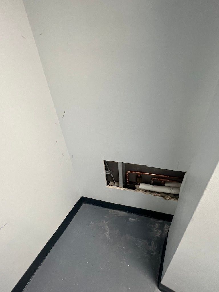 Small utility opening in a white wall above a gray floor, with exposed pipes visible inside.