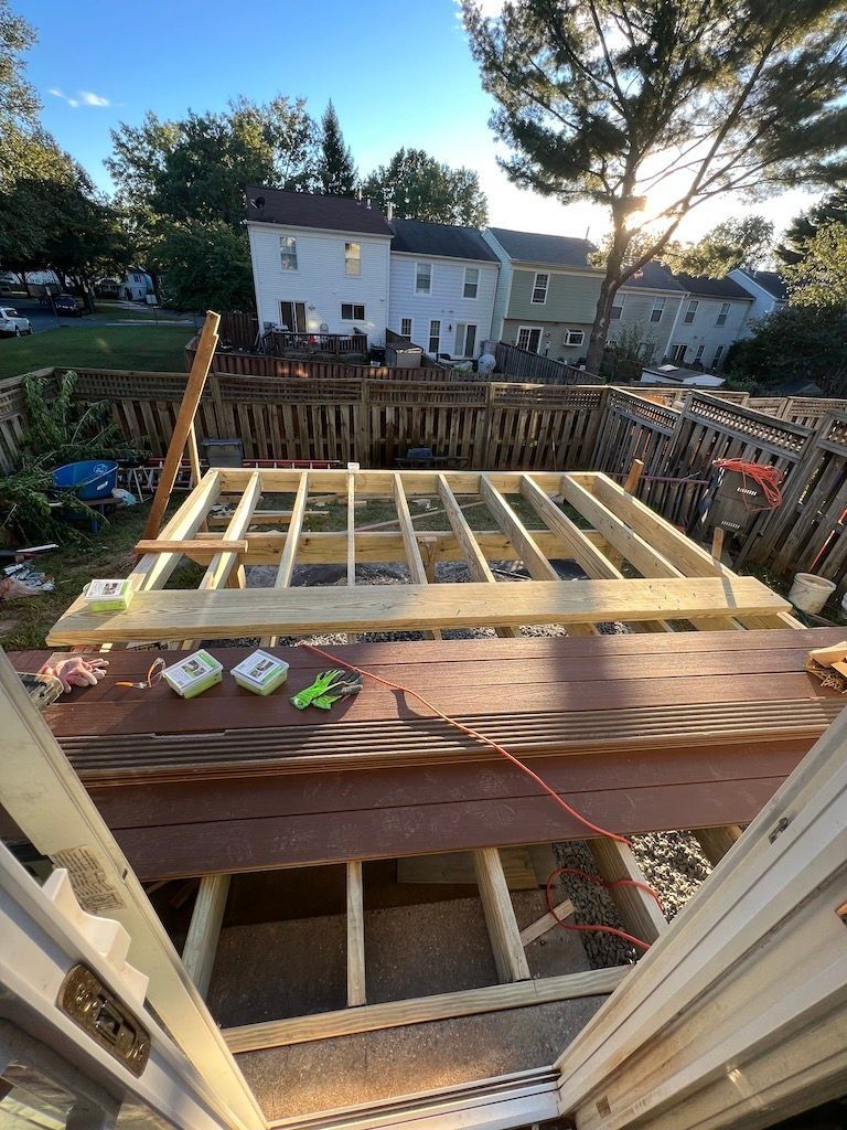 Wood-framed deck under construction, viewed from a doorway, with houses and trees in the background.