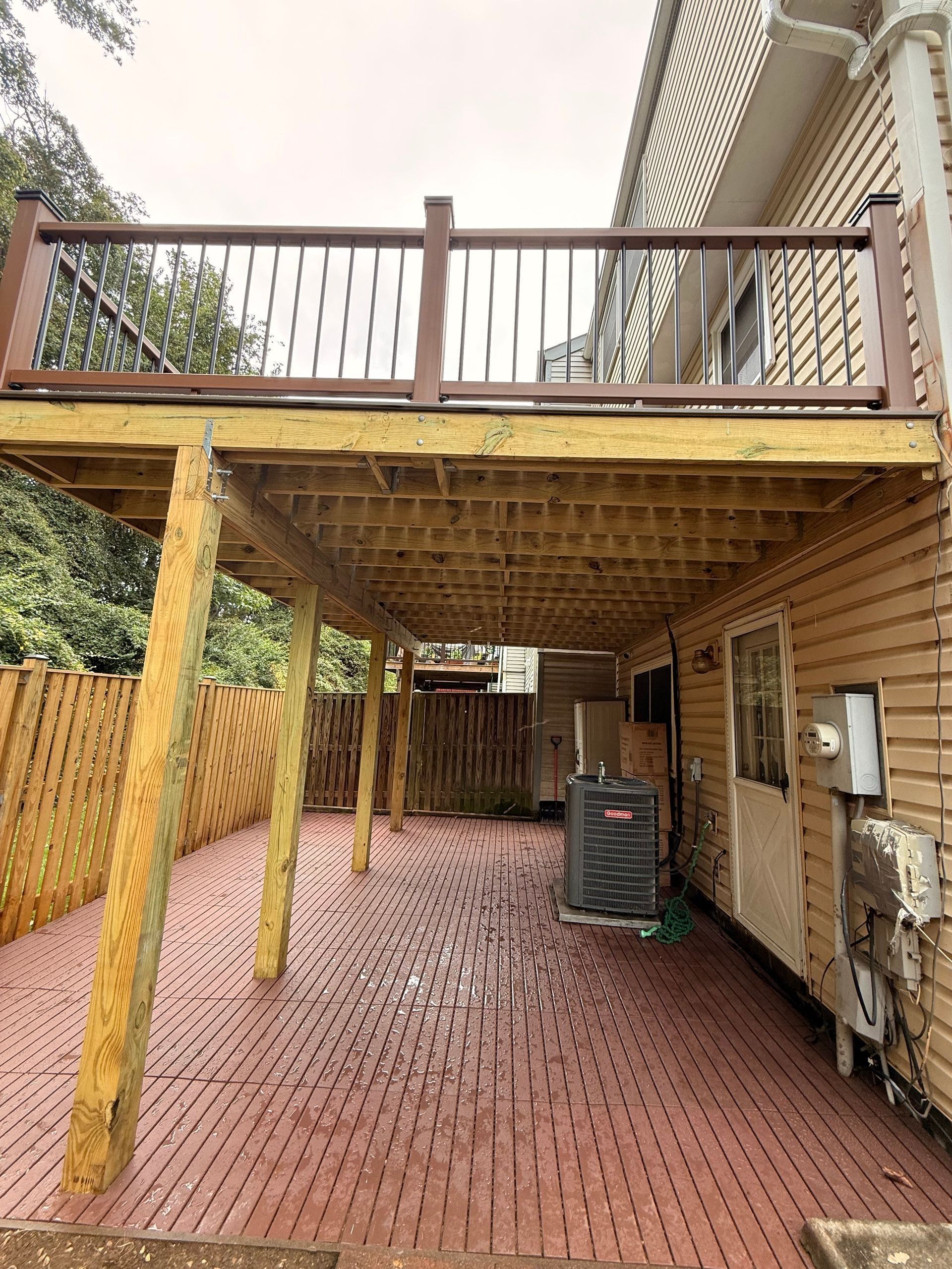Covered wooden deck over red brick patio beside house, with stairs and railing above.