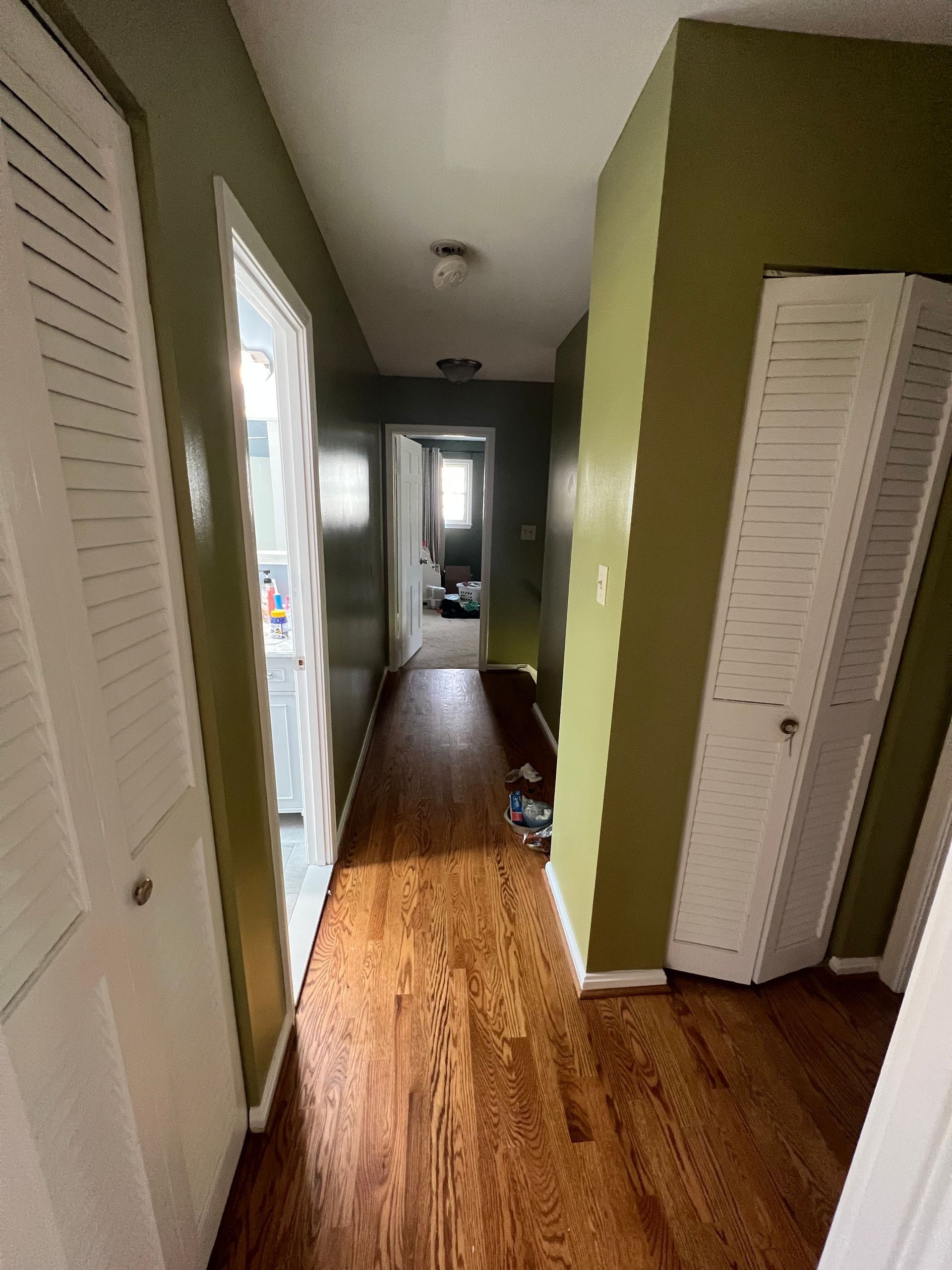 Narrow hallway with wood floors, green walls, white closet doors, and a bright doorway at the end