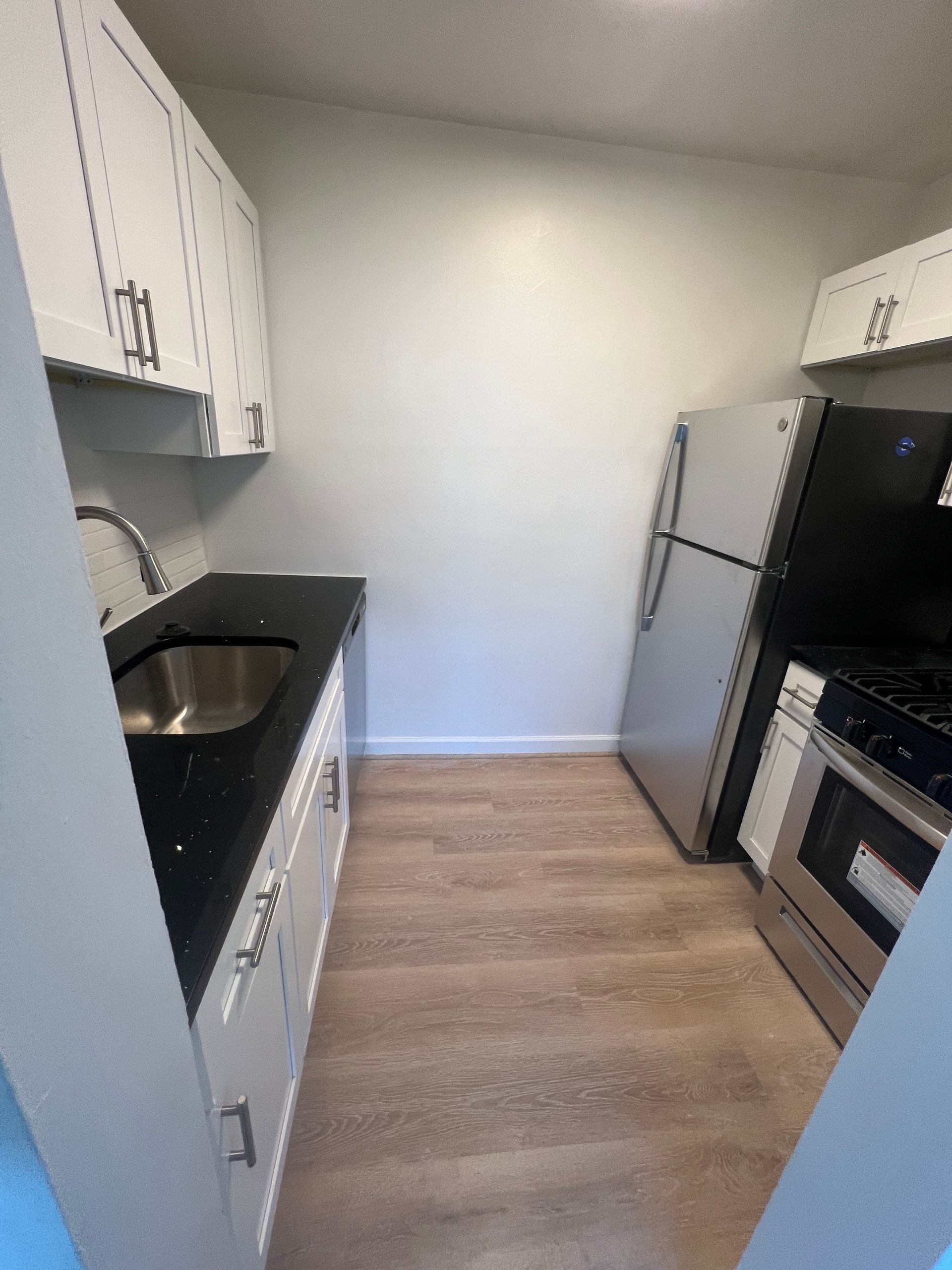 Small narrow kitchen with white cabinets, black countertops, stainless sink, and a refrigerator.