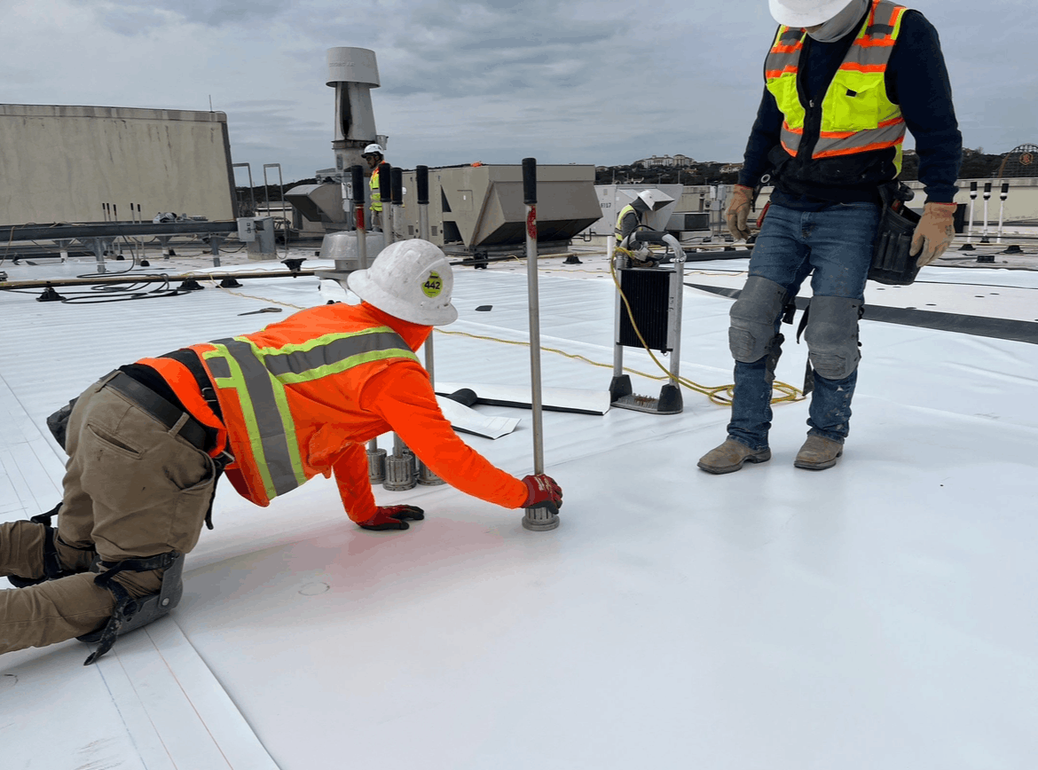 Worker in orange safety vest kneels on snowy rooftop, measuring near a standing colleague.