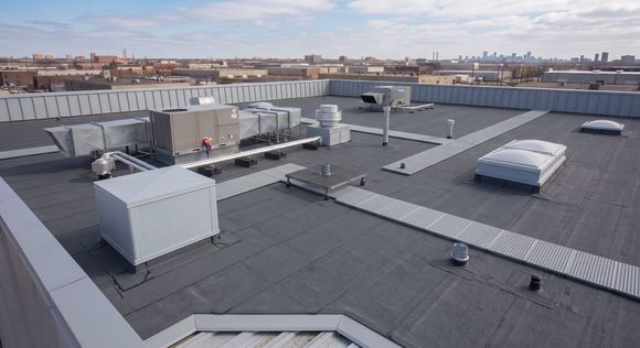 Rooftop view of flat gray roofs with vents and skylights under a cloudy sky, city skyline in the distance