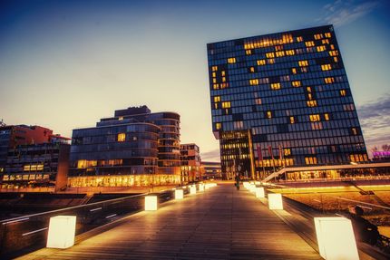 Modern waterfront buildings at dusk, lit walkway leading toward a tall illuminated tower under a blue sky