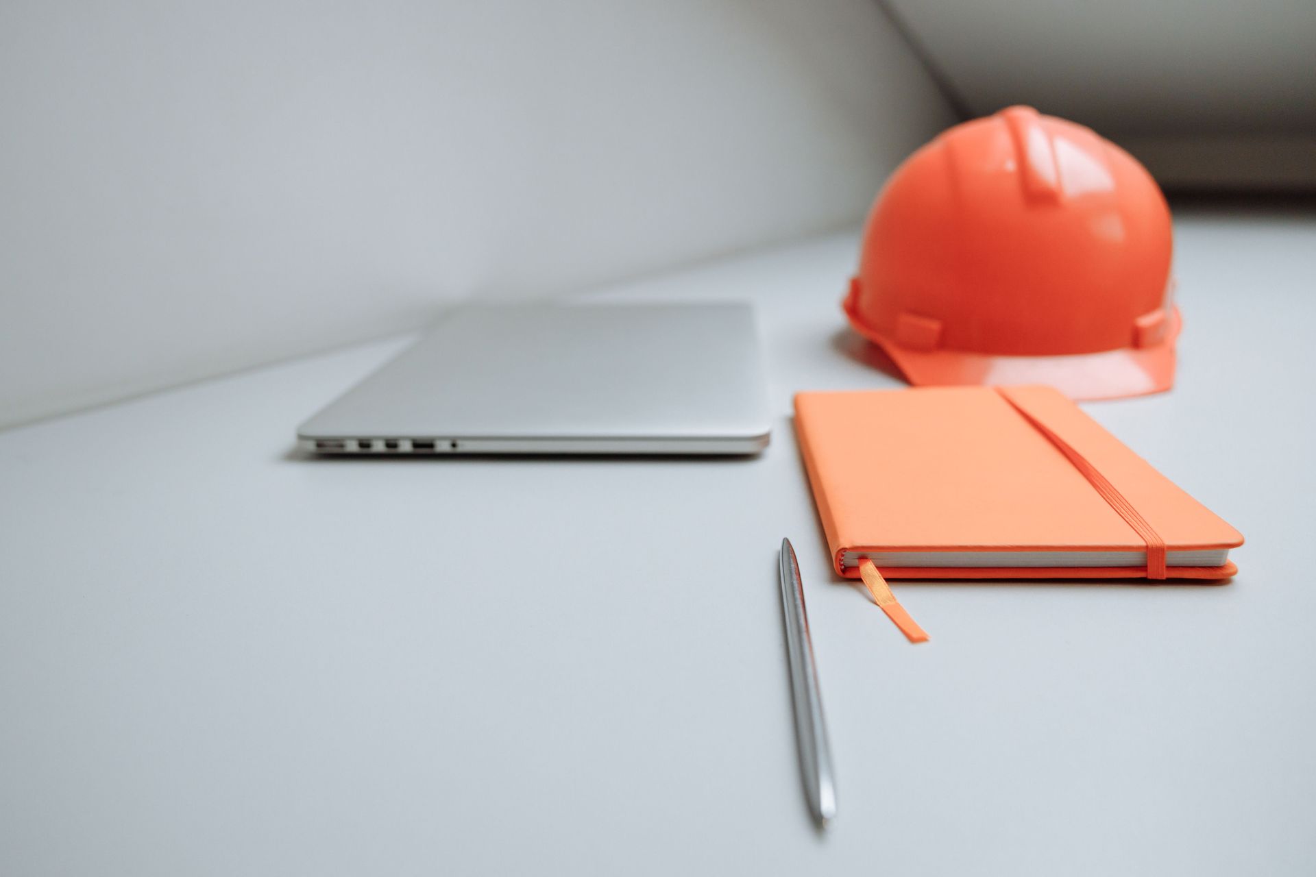 Silver laptop, orange hard hat, orange notebook, and pen on a white desk.