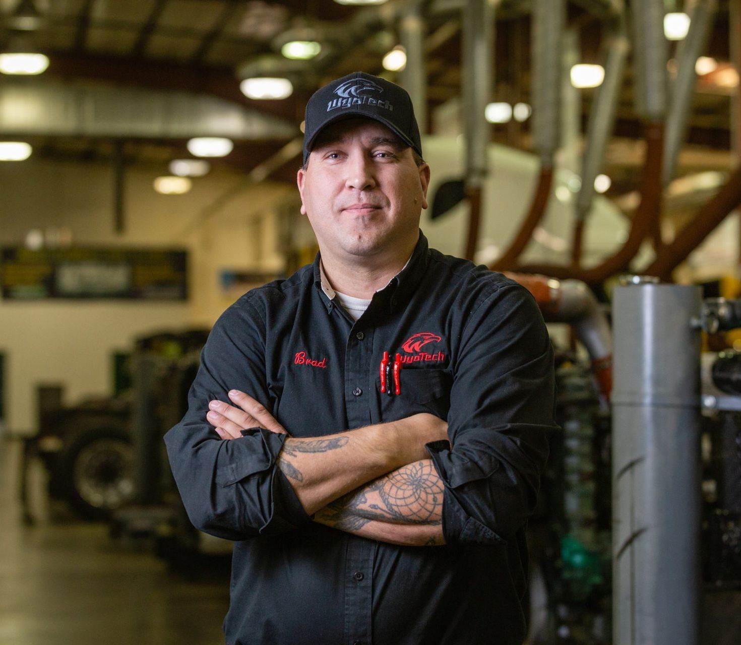 A man in a black shirt and hat is standing in a garage with his arms crossed.