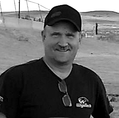 A man wearing a hat and sunglasses is smiling in a black and white photo.