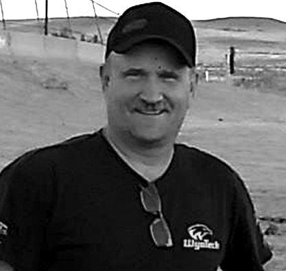 A man wearing a hat and sunglasses is smiling in a black and white photo.
