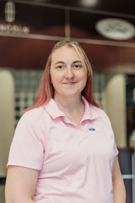 Woman with pink hair wearing a pink polo shirt, smiling in front of a Ford dealership logo.