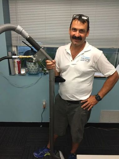Man holding a large hose in an indoor workspace, wearing a white polo and dark pants.