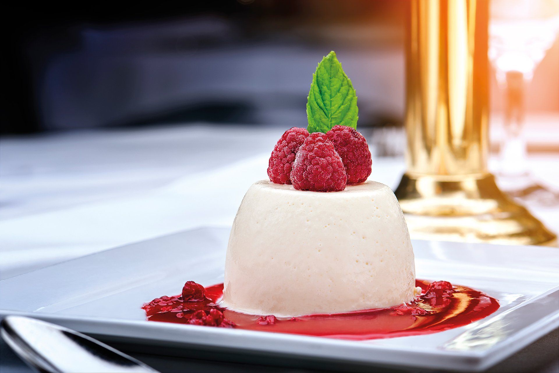 Panna cotta dessert with raspberries and mint on a white plate, with a golden object and blurred background.