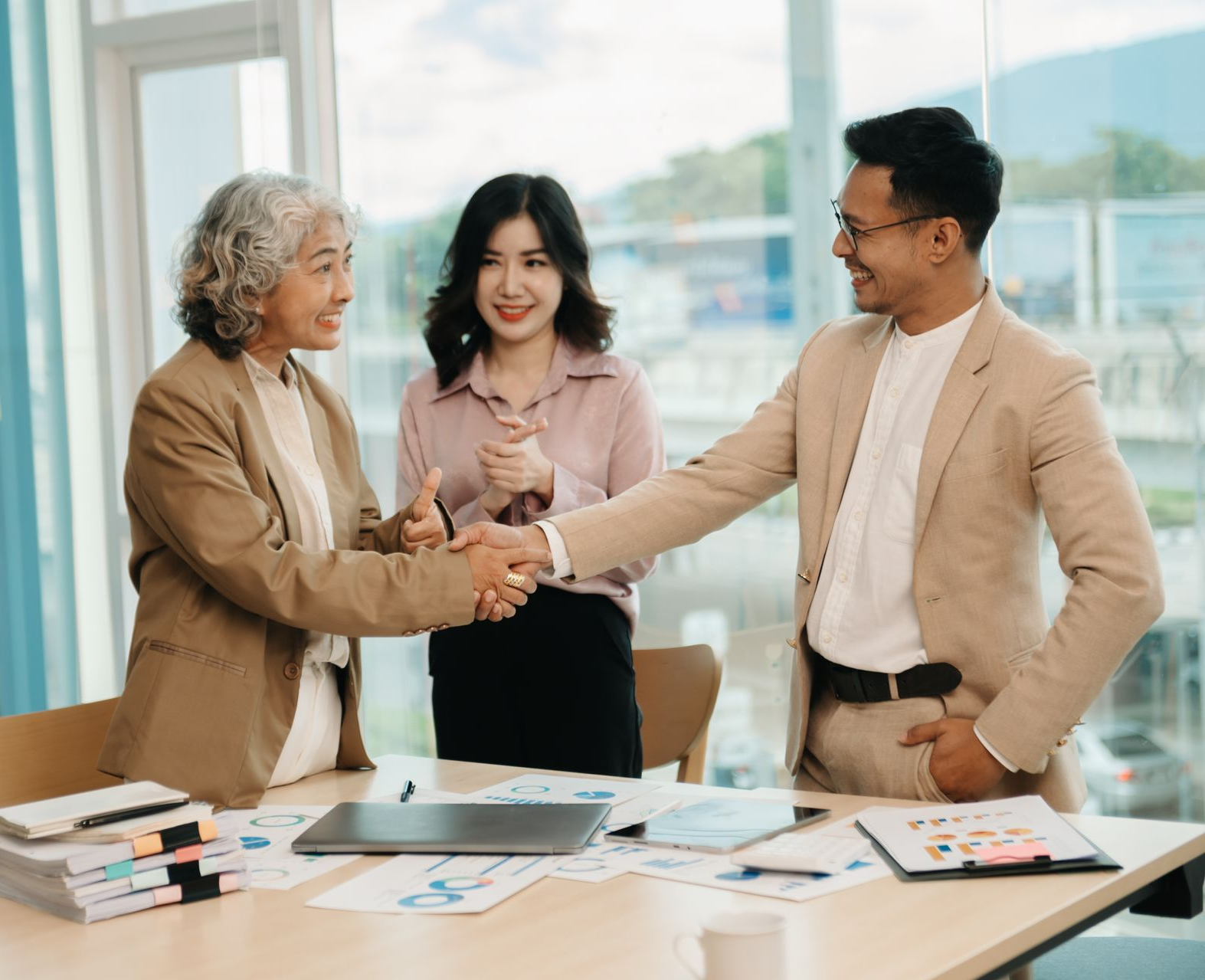 Businesspeople shaking hands, celebrating a deal at a desk, office setting. Woman in pink smiles.