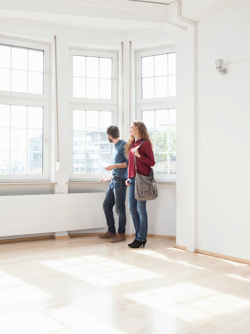 Couple looking out of large windows in an empty room with light wood floors.