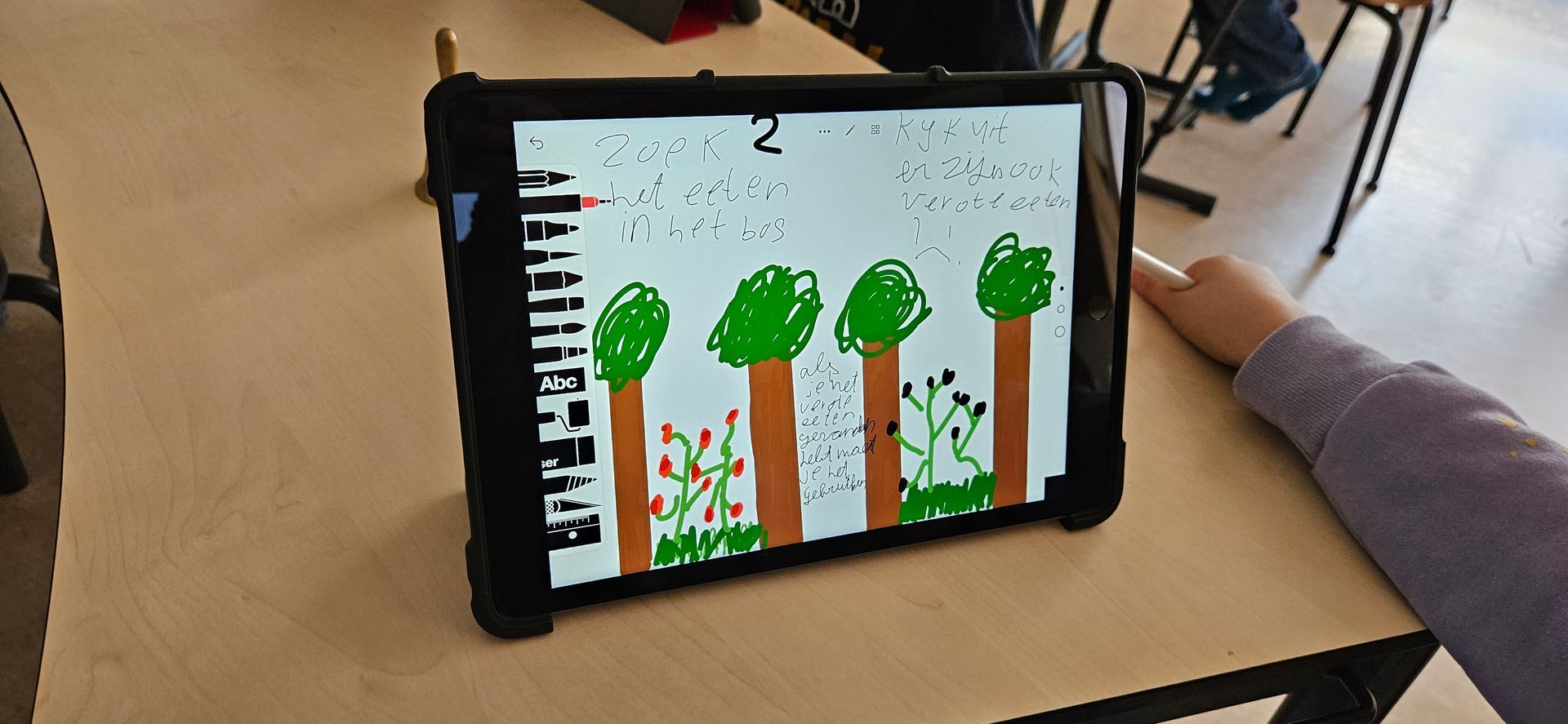 Een tekening van bomen op een tabletscherm, dat op een licht houten bureau staat, met een zichtbare hand.