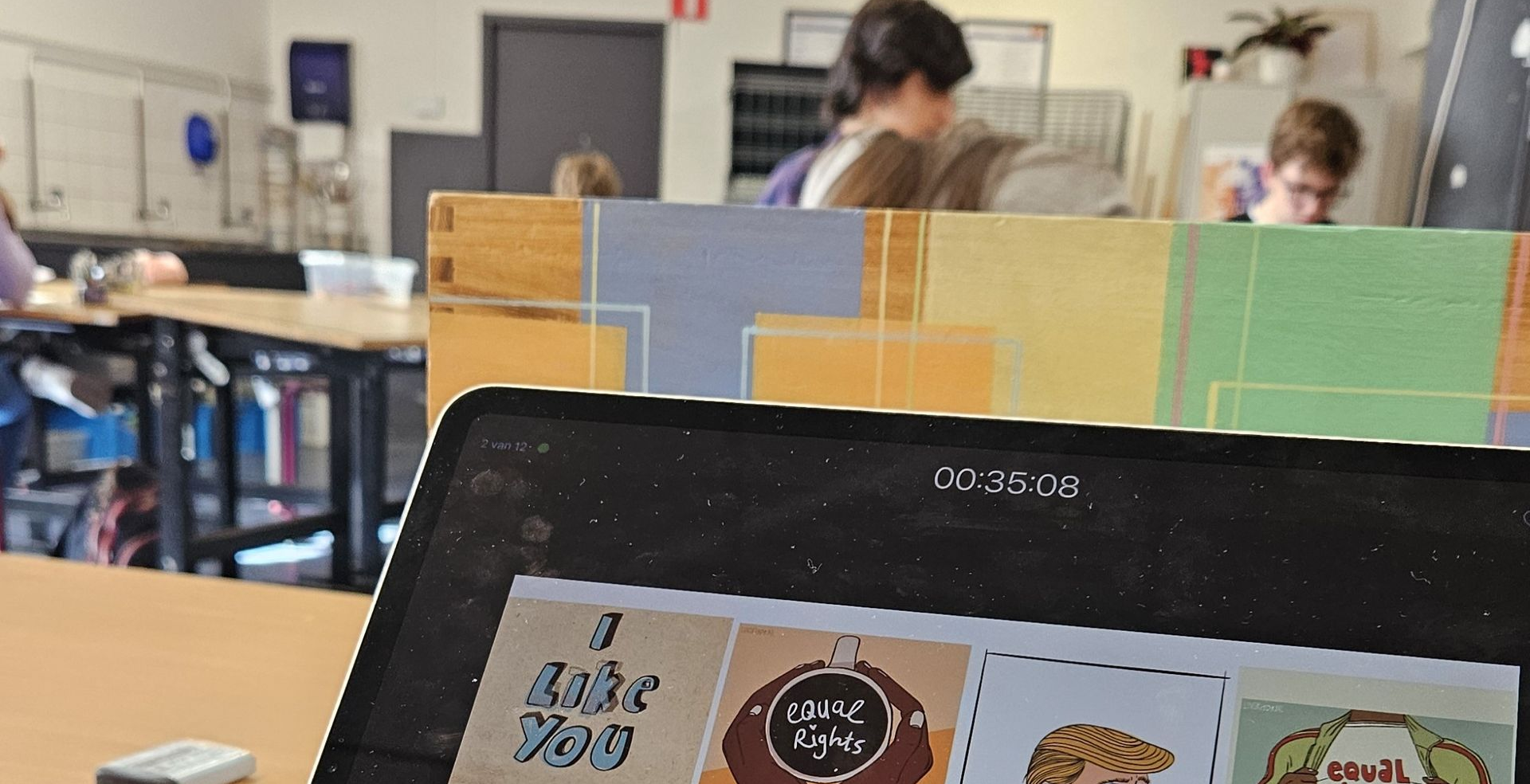 Een laptopscherm met kleurrijke kaarten in een klaslokaal; op de achtergrond zitten mensen aan bureaus.