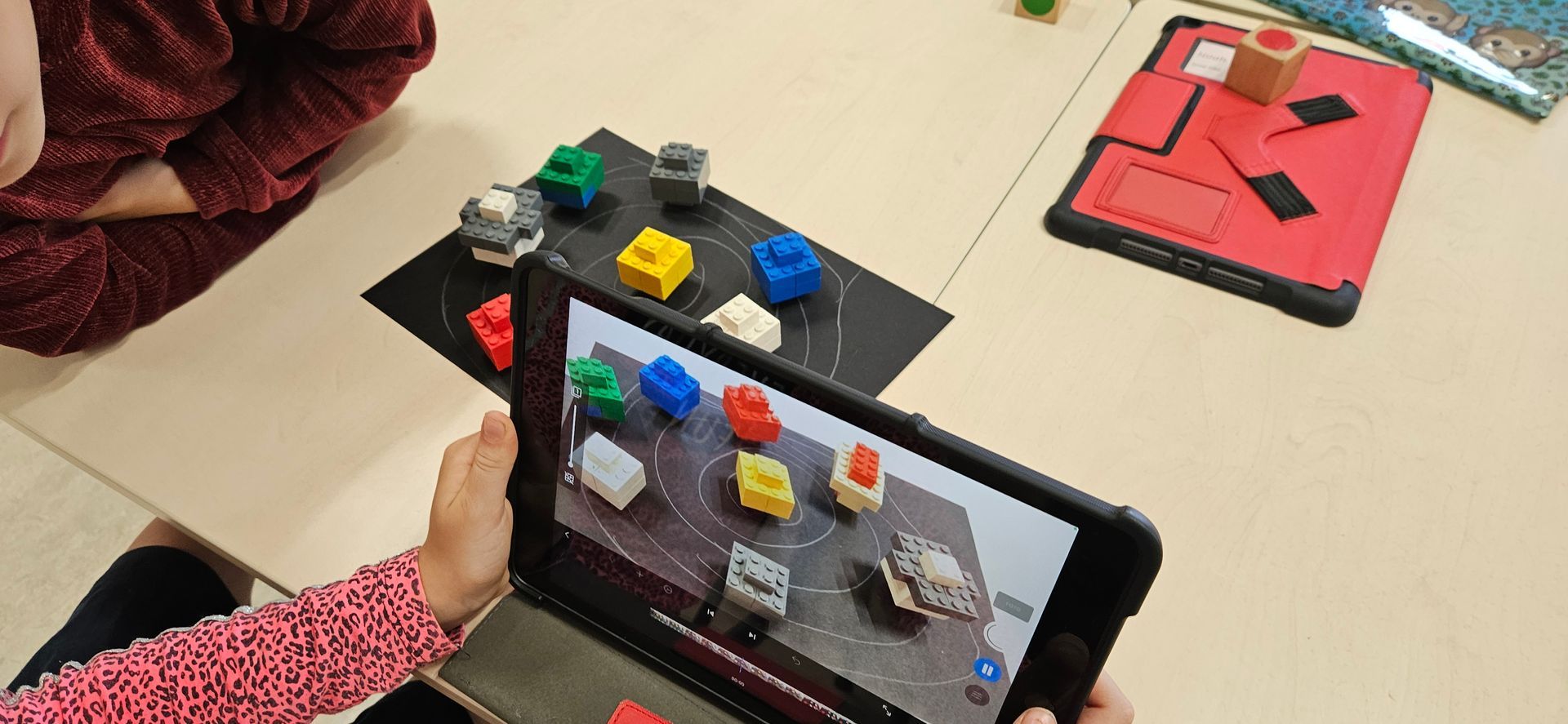 Een kind houdt een tablet vast waarop kleurrijke blokken op een tafel worden weergegeven, mogelijk met behulp van augmented reality.