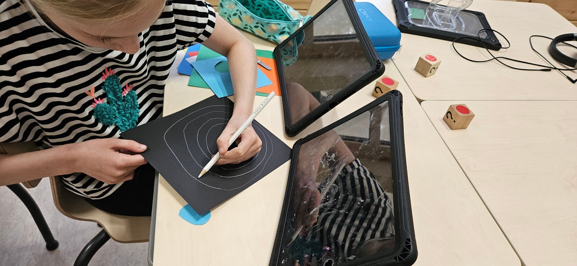 Een persoon tekent met een witte pen op zwart papier aan een tafel. Tablets en gekleurd papier liggen in de buurt.