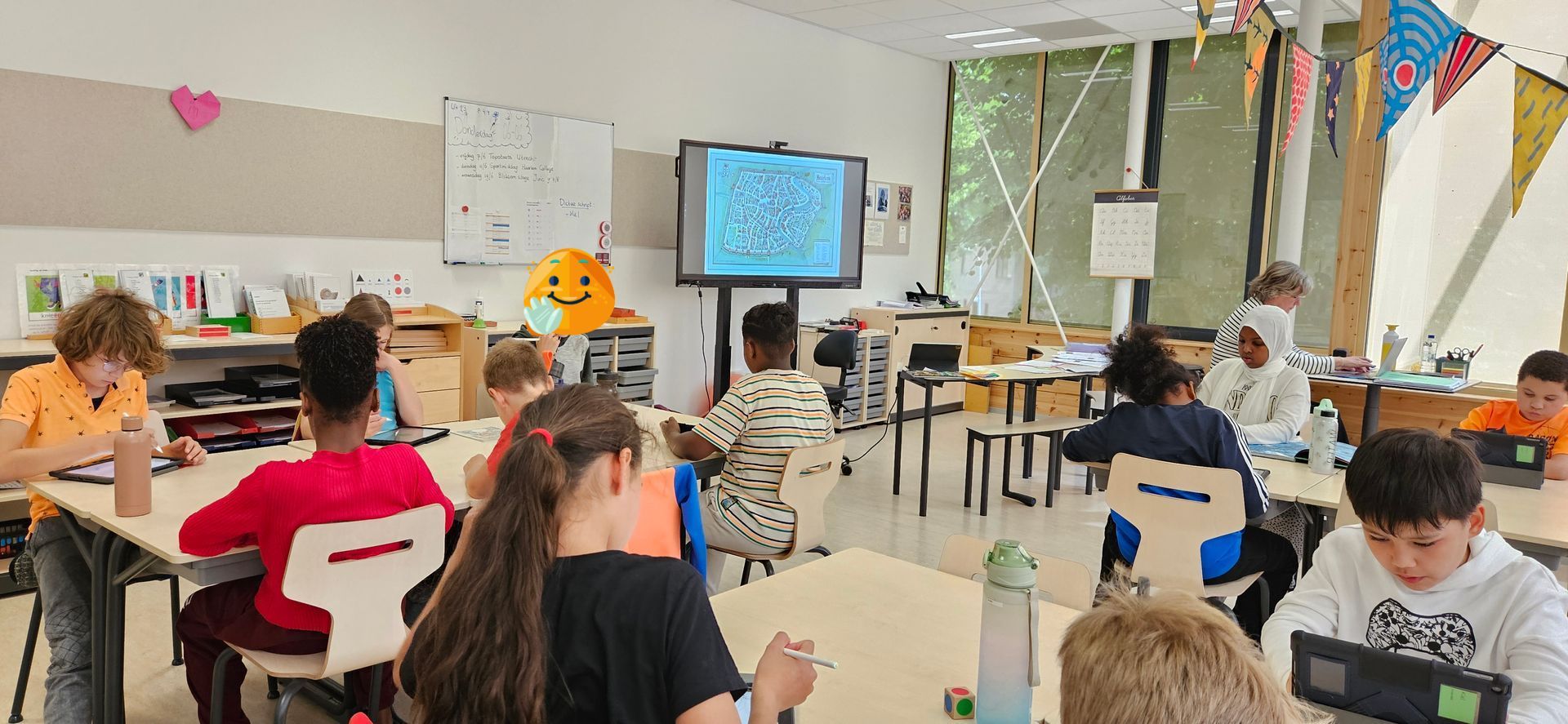 Studenten werken op laptops aan bureaus in een klaslokaal met een groot scherm waarop informatie wordt weergegeven.