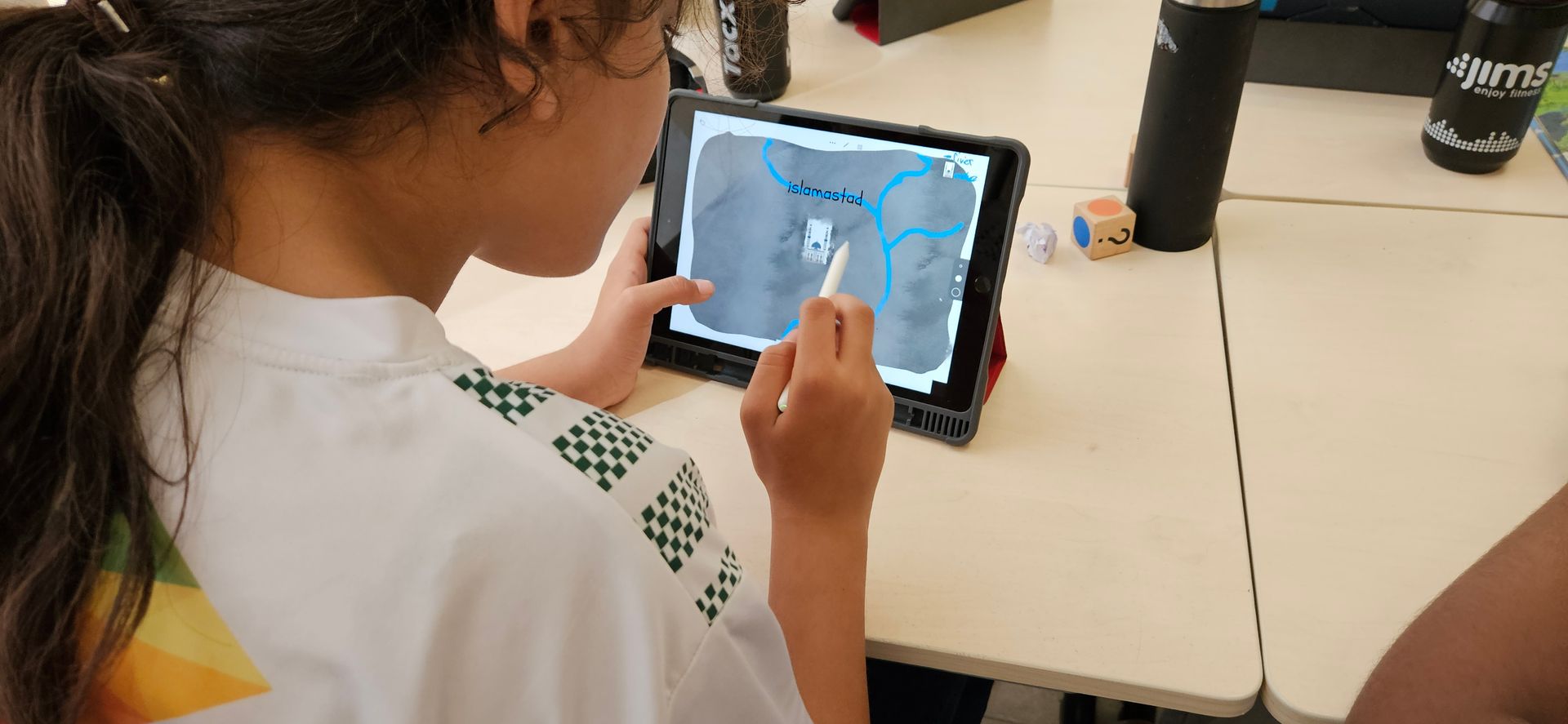 Een leerling gebruikt een stylus op een tablet om over een kaart te tekenen, aan een bureau in een klaslokaal.