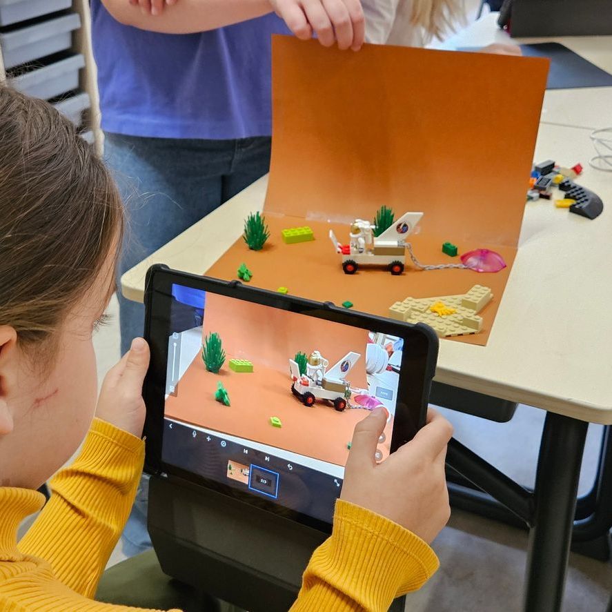 Een kind gebruikt een tablet om een ​​Lego-scène te filmen met een klein voertuig en bomen tegen een oranje achtergrond.