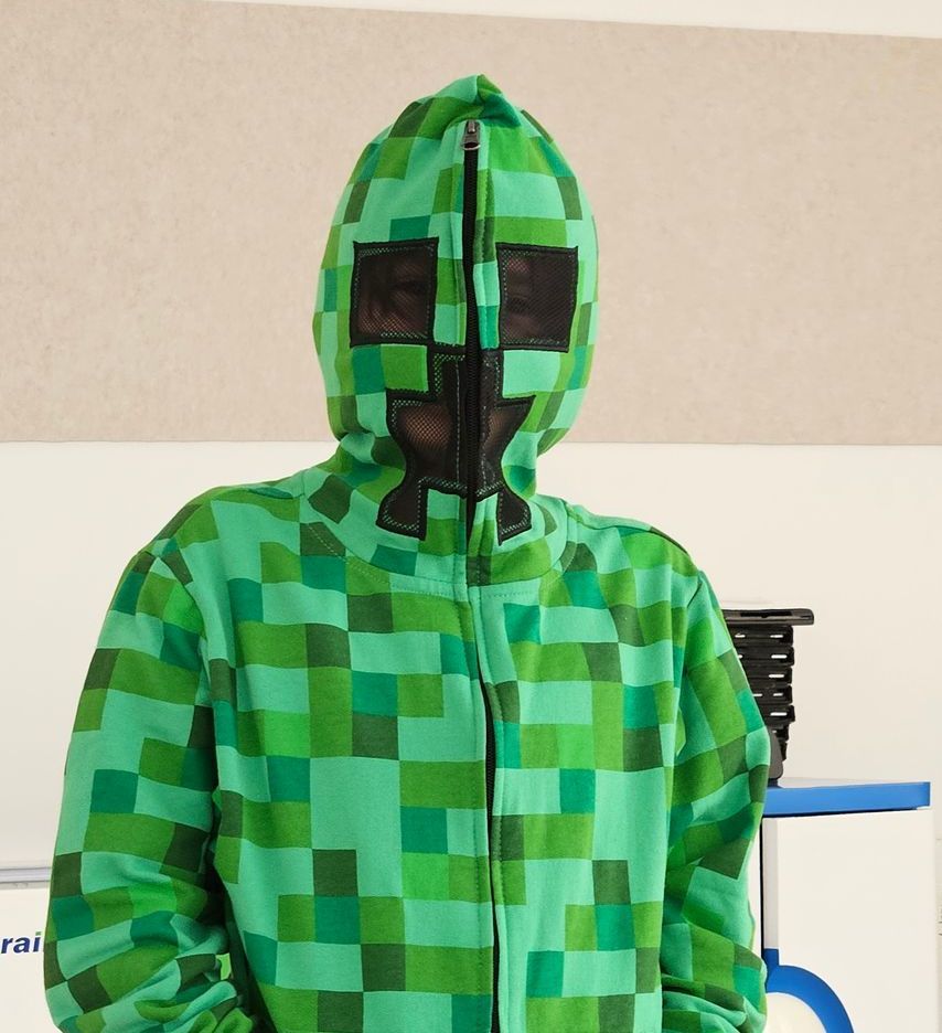 Persoon in een groene, Creeper-achtige Minecraft-jumpsuit met capuchon, waarvan de rits open is, staat binnen.