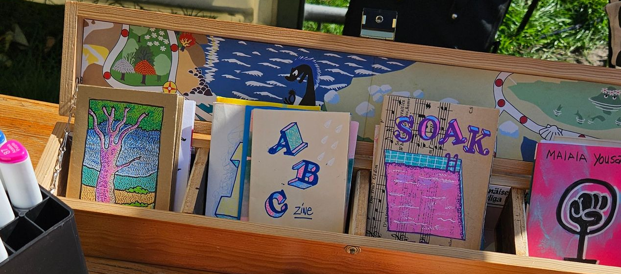 Kunstwerken tentoongesteld in een houten rek; kleurrijke tekeningen, waaronder de letters ABC en een tekening van een boom, zijn zichtbaar.