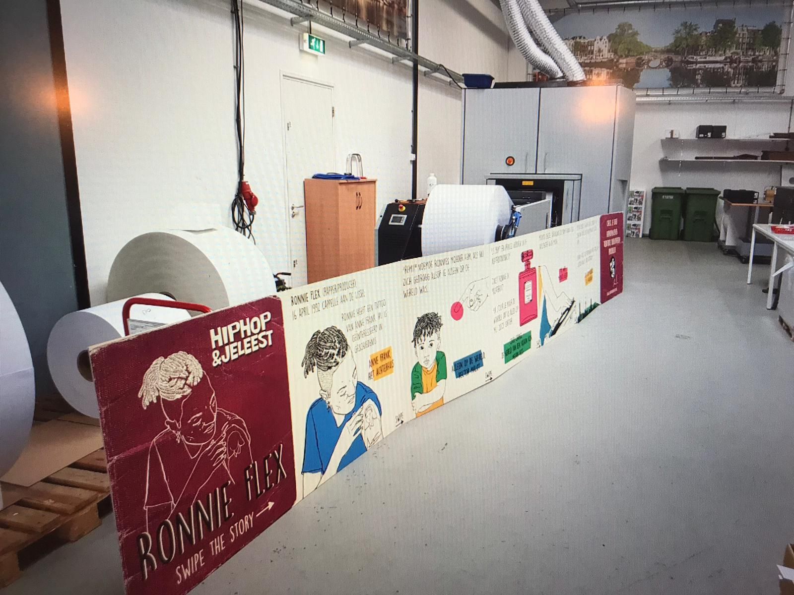 Drukpers met papierrollen en een lange grafische weergave met illustraties in een industriële omgeving.