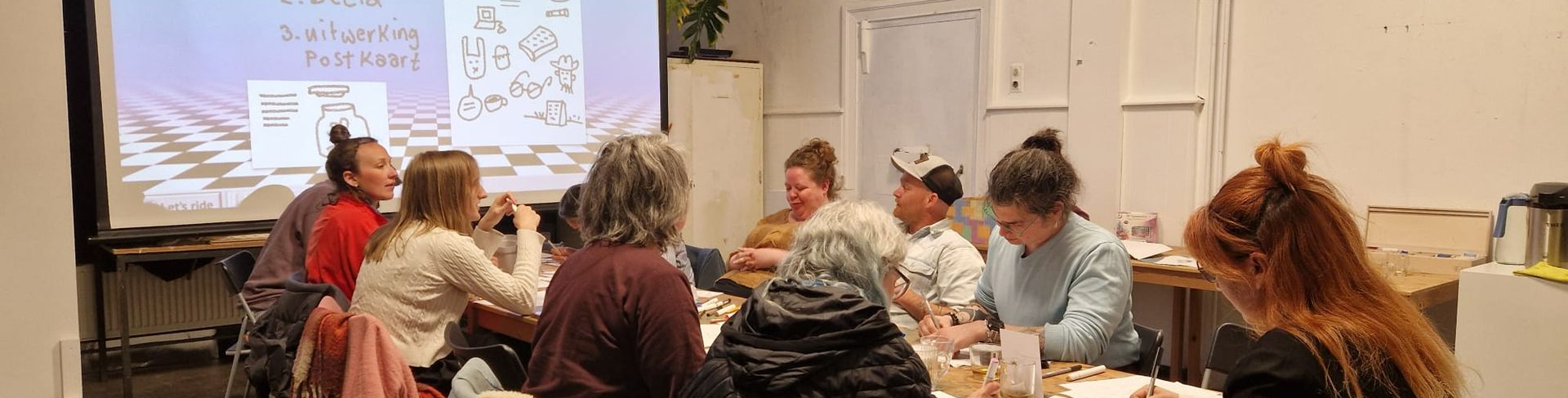 Een groep mensen zit aan een tafel in een kamer en kijkt naar beneden. Op de achtergrond is een projectiescherm te zien.