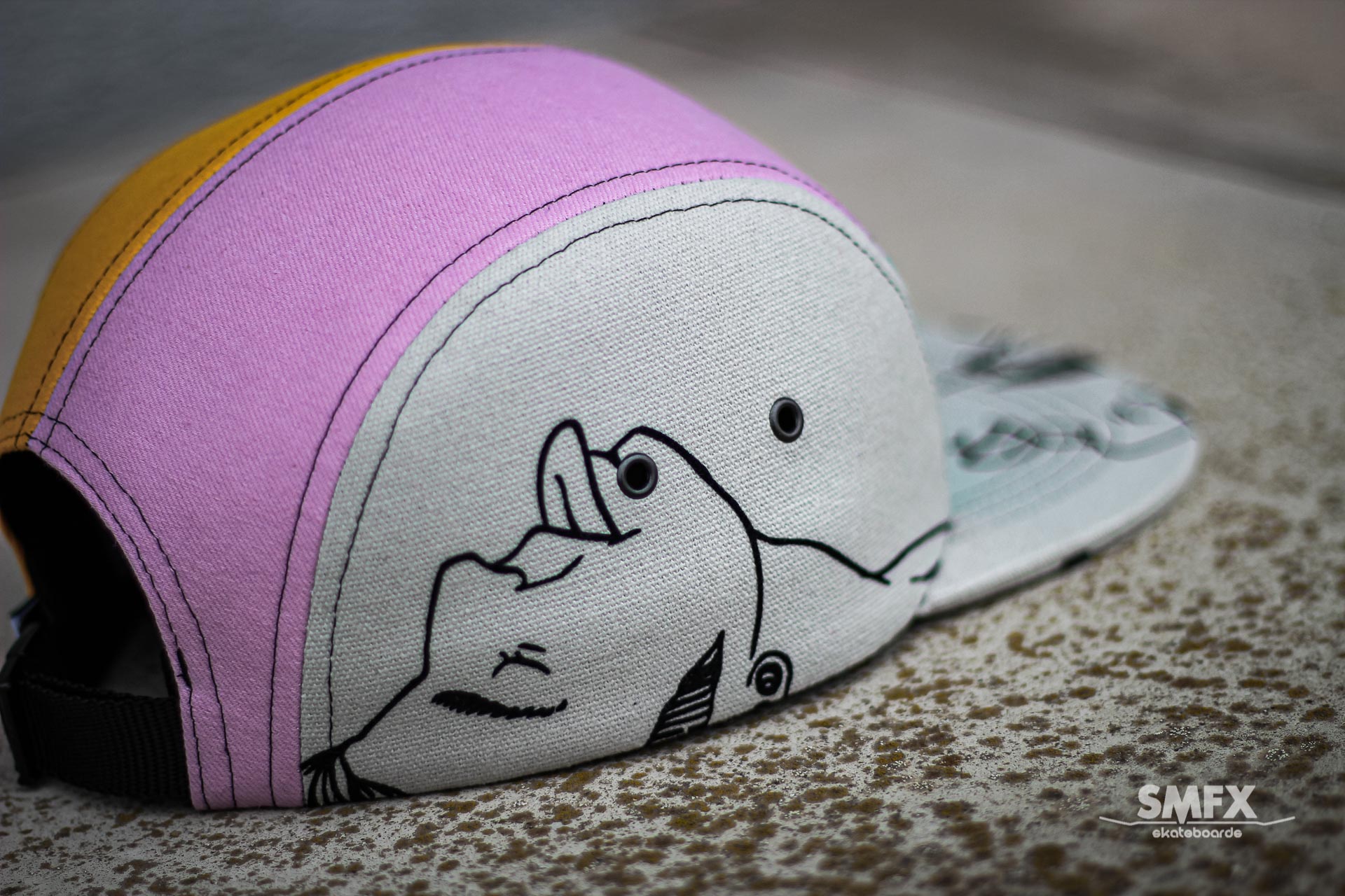 Close-up van een kleurrijke pet met een cartoongezichtje aan de zijkant. Roze, witte en gele panelen met een zwarte omlijning.