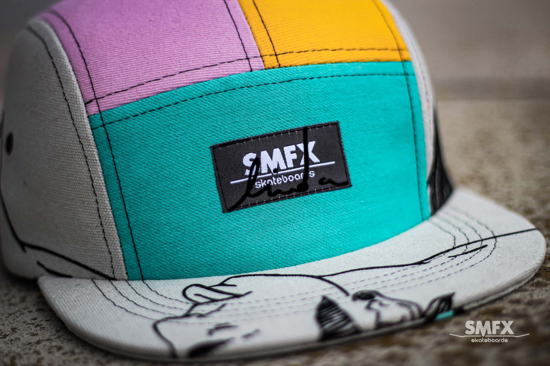 Een kleurrijke pet met vijf panelen en accenten in roze, geel en turquoise. De klep heeft een zwart-wit design met het SMFX-logo.