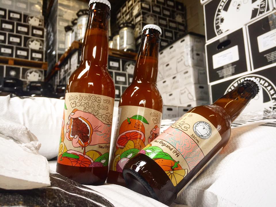 Drie bierflessen met kleurrijke fruitetiketten; de flessen staan ​​op een wit kleed, met dozen en schappen op de achtergrond.