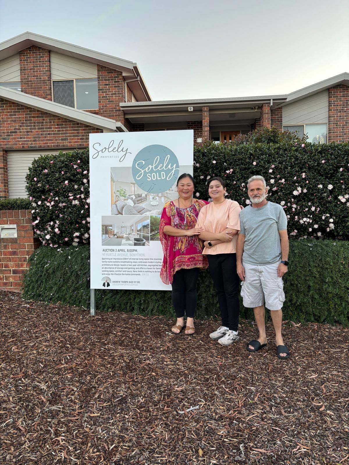 Three people standing beside a sign in front of a brick building and hedges, smiling outdoors