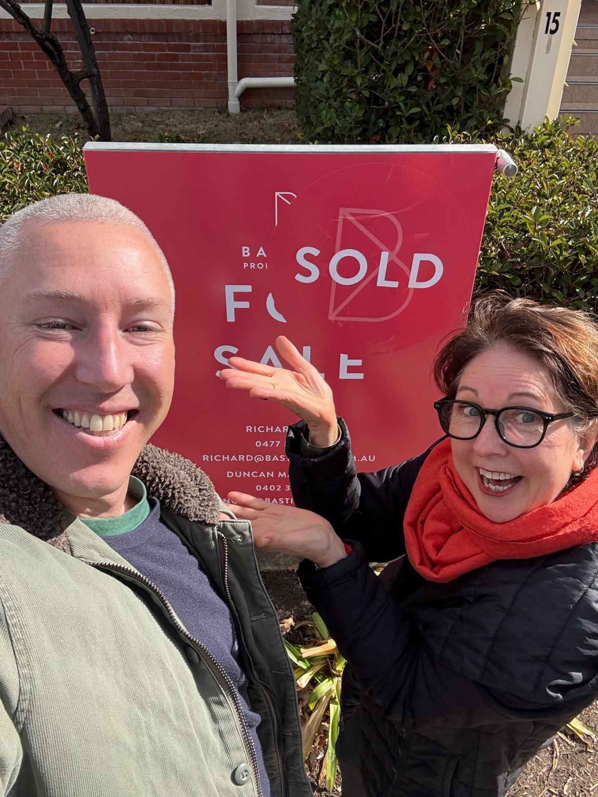 Two smiling people posing beside a red “SOLD” sign outdoors