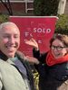 Two smiling people posing beside a red “SOLD” sign outdoors