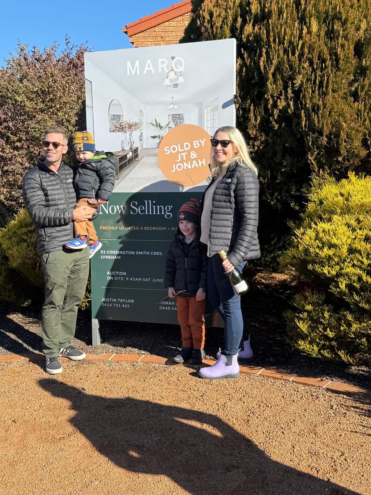 Family posing outdoors beside a real estate sign on a sunny day