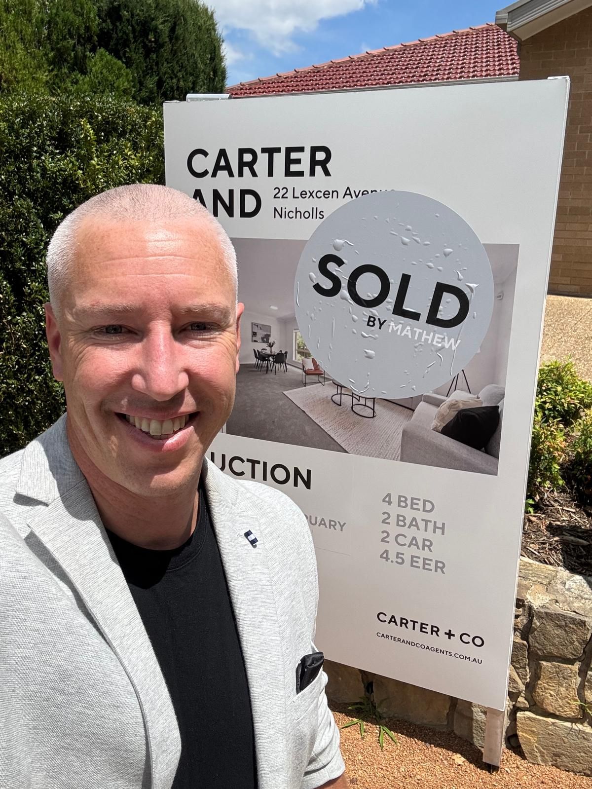 Smiling man stands beside a real estate sign with a large “SOLD” notice.